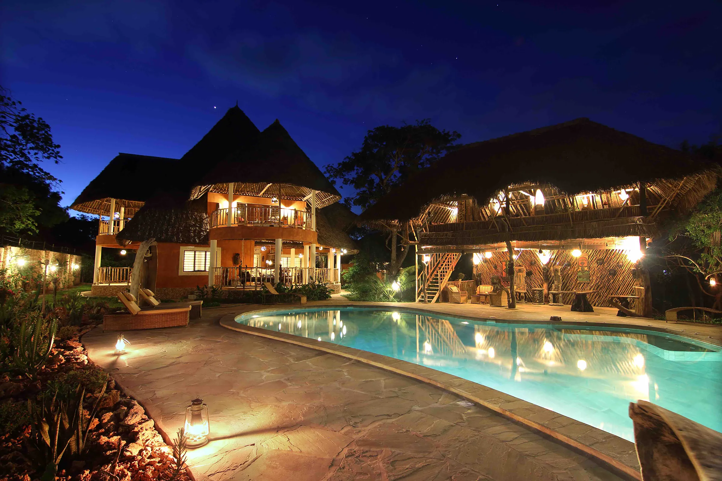 The villa lit at night, makuti roofs above the pool